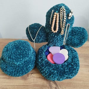 May include: Teal crochet jewelry organizer shaped like a cactus, with a separate round container. The cactus holds necklaces and earrings. The open compartment contains colorful heart-shaped objects. The organizer sits on a wooden surface.