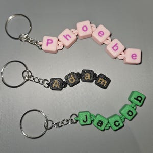 May include: Three keychains with different names spelled out in colourful beads. The first keychain is pink with the name "Phoebe" spelled out. The second keychain is black with the name "Adam" spelled out. The third keychain is green with the name "Jacob" spelled out.
