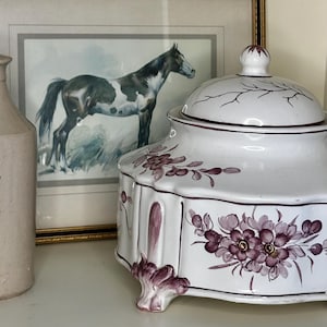 May include: A beige ceramic bottle, a white ceramic lidded jar with purple floral patterns and scalloped edges, and a framed watercolour painting of a black and white horse standing in a field, all arranged together on a flat surface.