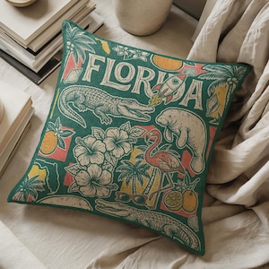 May include: Square green decorative pillow with a Florida theme. The pillow features illustrations of an alligator, manatee, flamingo, hibiscus flowers, and the word "FLORIDA" in white. Accents include oranges, palm trees, and a rocket ship.