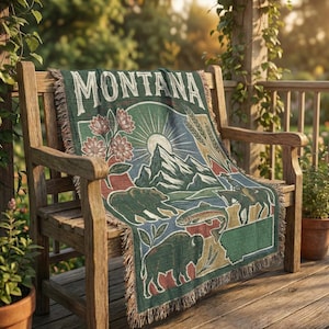 May include: A woven throw blanket with a Montana-themed design, draped over a wooden bench. The blanket features illustrations of mountains, a bear, a bison, and the text "MONTANA" in a vintage style. The color palette includes green, blue, and tan.