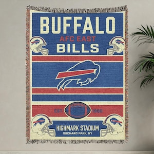 May include: A blue, red, and cream-colored woven throw blanket featuring the text "BUFFALO AFC EAST BILLS." The blanket includes the team logo, football helmets, and a football. The text "HIGHMARK STADIUM ORCHARD PARK, NY" is also visible.