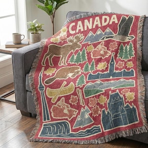 May include: A red throw blanket with a fringe edge, featuring various Canadian icons such as a moose, mountains, and maple leaves. The word "CANADA" is displayed across the top in cream-colored letters. The blanket is draped over a gray sofa.