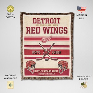 May include: A woven cotton throw blanket featuring the Detroit Red Wings logo, text, and hockey imagery. The design includes crossed hockey sticks, a puck, two helmets, and the text Detroit Red Wings, Atlantic Division, Est. 1926, Little Caesars Arena.