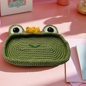 May include: A green crocheted frog-shaped tray with black and white eyes and a yellow crown. The tray is on a pink surface. The tray is a decorative item.