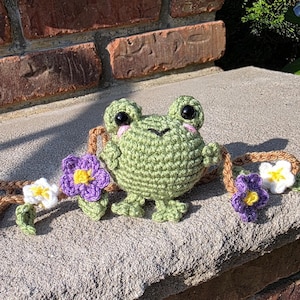 May include: A handmade green crochet frog with large black eyes and pink cheeks. The frog is adorned with a brown vine featuring purple and white crochet flowers. The frog is sitting on a stone surface.