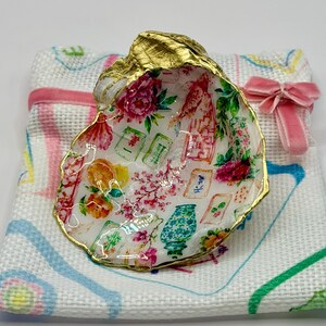 K&ouml;nnte beinhalten: Dekorative Austernschale mit goldfarbenem Rand, verziert mit einem floralen und asiatisch inspirierten Design. Sie liegt auf einem wei&szlig;en Tuch mit einer rosa Schleife. Das Innere der Schale zeigt ein farbenfrohes Muster aus Blumen und asiatischen Motiven.