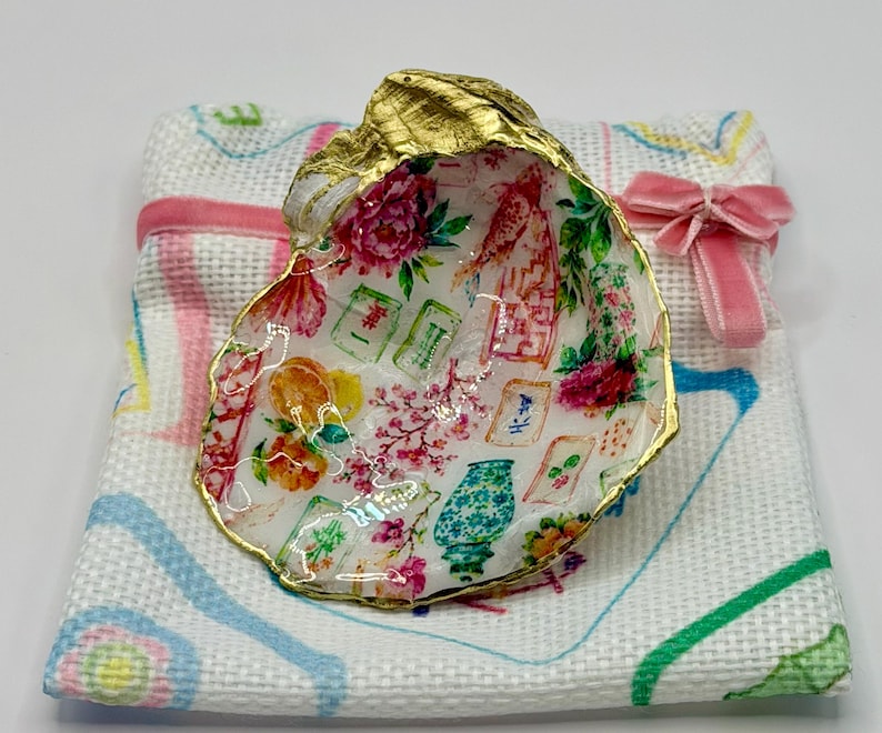 K&ouml;nnte beinhalten: Dekorative Austernschale mit goldfarbenem Rand, verziert mit einem floralen und asiatisch inspirierten Design. Sie liegt auf einem wei&szlig;en Tuch mit einer rosa Schleife. Das Innere der Schale zeigt ein farbenfrohes Muster aus Blumen und asiatischen Motiven.