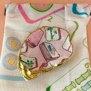 Puede incluir: Un plato decorativo de concha de ostra con un patr&oacute;n de decoupage de fichas de mahjong en rosa, blanco y verde, con un borde pintado en oro met&aacute;lico, sobre una bolsa de tela blanca con cord&oacute;n y un dise&ntilde;o de cuadr&iacute;cula impreso.