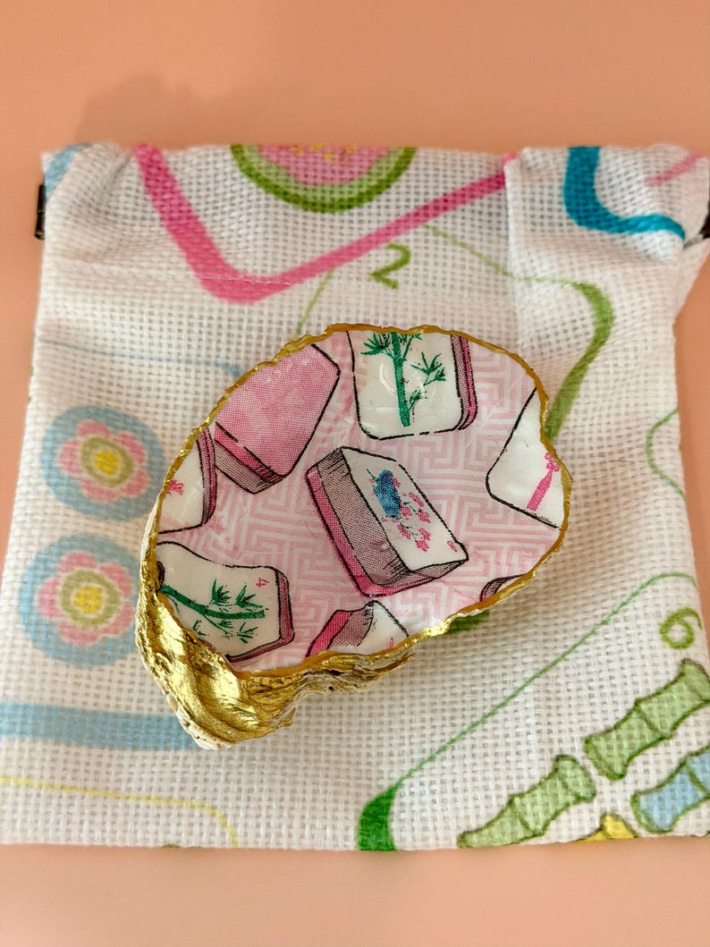 Puede incluir: Un plato decorativo de concha de ostra con un patr&oacute;n de decoupage de fichas de mahjong en rosa, blanco y verde, con un borde pintado en oro met&aacute;lico, sobre una bolsa de tela blanca con cord&oacute;n y un dise&ntilde;o de cuadr&iacute;cula impreso.