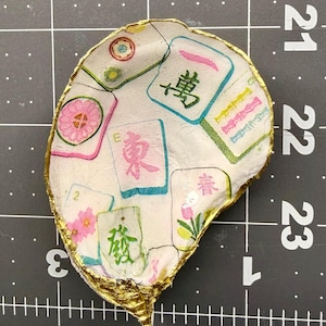 K&ouml;nnte beinhalten: Eine handgefertigte, muschelf&ouml;rmige Schmuckschale. Die Schale ist wei&szlig; mit einem Muster aus bunten Mahjong-Steinen. Die R&auml;nder sind mit goldener Farbe verziert. Die Schale befindet sich auf einer schwarzen Rasteroberfl&auml;che.