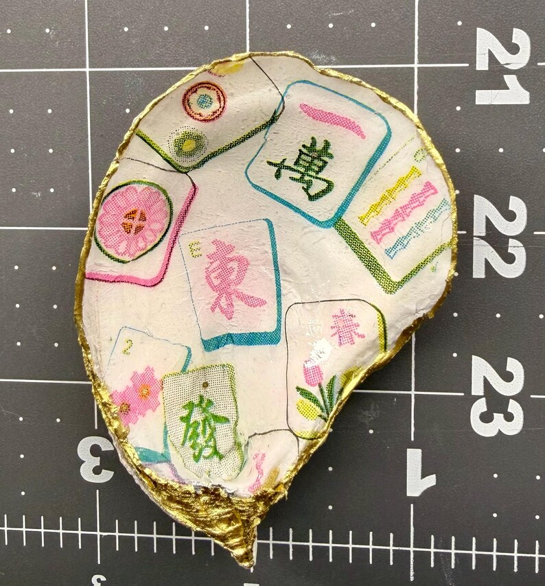 K&ouml;nnte beinhalten: Eine handgefertigte, muschelf&ouml;rmige Schmuckschale. Die Schale ist wei&szlig; mit einem Muster aus bunten Mahjong-Steinen. Die R&auml;nder sind mit goldener Farbe verziert. Die Schale befindet sich auf einer schwarzen Rasteroberfl&auml;che.