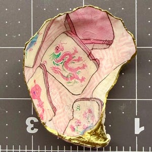 Puede incluir: Un plato decorativo hecho de concha de ostra con un patr&oacute;n de decoupage de fichas de mahjong rosas y blancas, incluyendo una ficha de drag&oacute;n rosa. La concha tiene un borde festoneado pintado con un acabado dorado met&aacute;lico sobre una rejilla.
