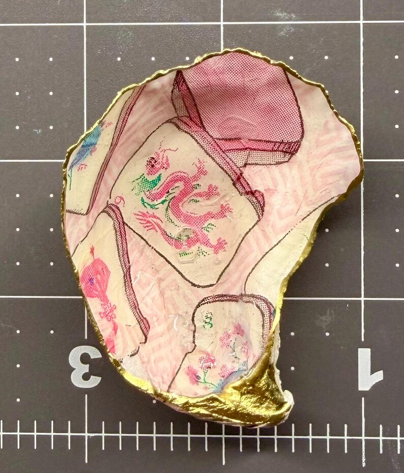 Puede incluir: Un plato decorativo hecho de concha de ostra con un patr&oacute;n de decoupage de fichas de mahjong rosas y blancas, incluyendo una ficha de drag&oacute;n rosa. La concha tiene un borde festoneado pintado con un acabado dorado met&aacute;lico sobre una rejilla.