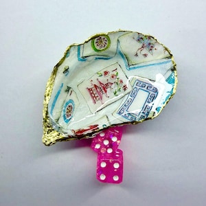 K&ouml;nnte beinhalten: Ein Muschelschalen-Schmucksch&auml;lchen mit goldfarbenem Rand, verziert mit einem Vintage-Spielkartenmuster. Drei rosa W&uuml;rfel mit wei&szlig;en Punkten befinden sich darunter. Ein einzigartiger Dekorationsartikel.