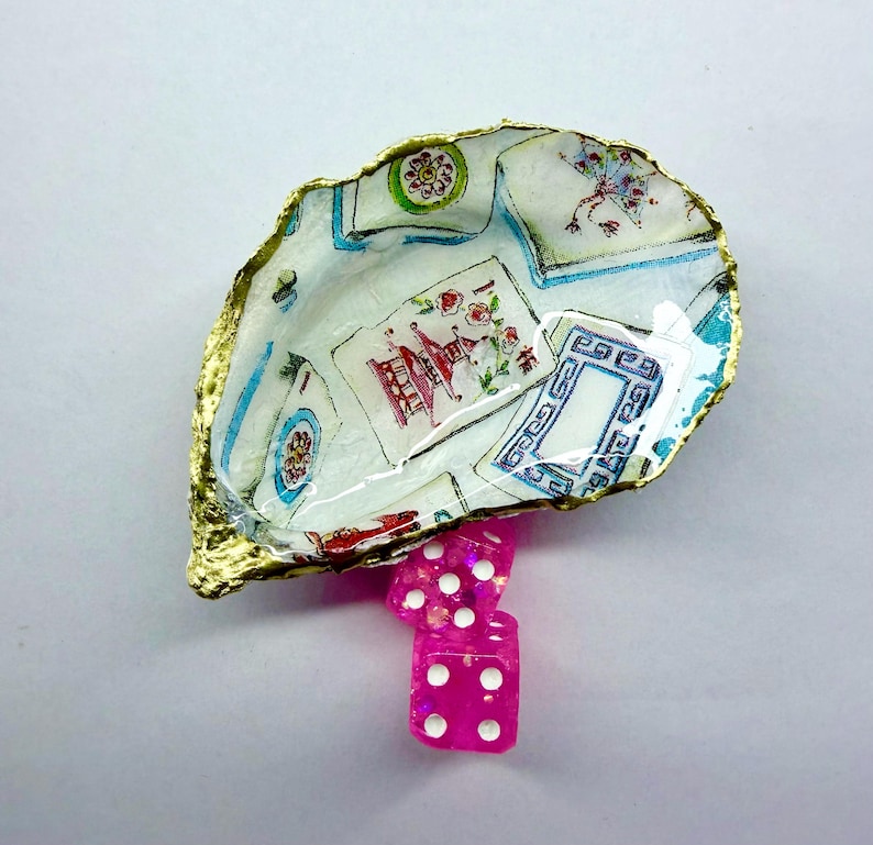 K&ouml;nnte beinhalten: Ein Muschelschalen-Schmucksch&auml;lchen mit goldfarbenem Rand, verziert mit einem Vintage-Spielkartenmuster. Drei rosa W&uuml;rfel mit wei&szlig;en Punkten befinden sich darunter. Ein einzigartiger Dekorationsartikel.