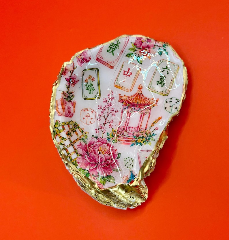 K&ouml;nnte beinhalten: Eine Austernschale, verziert mit einem farbenfrohen Design mit einer Pagode, Blumen, W&uuml;rfeln und Mahjong-Steinen. Die Schale hat einen goldfarbenen Rand und ist vor einem orangefarbenen Hintergrund platziert.