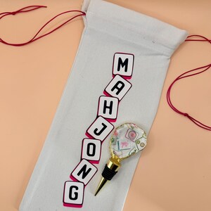 Puede incluir: Una bolsa de vino de lona blanca con la palabra "MAHJONG" impresa en negro y rosa. Un tap&oacute;n de botella decorativo con base dorada y parte superior transparente con estampado floral est&aacute; junto a la bolsa. Cierre de cord&oacute;n rojo.