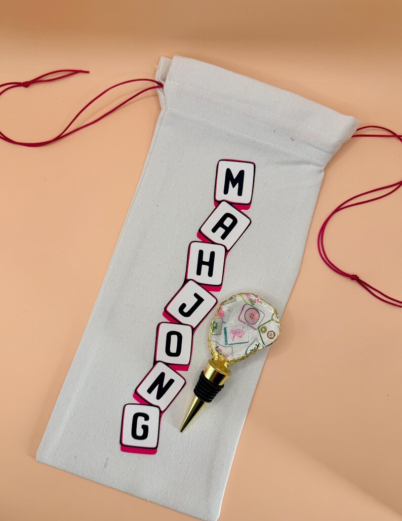 Puede incluir: Una bolsa de vino de lona blanca con la palabra "MAHJONG" impresa en negro y rosa. Un tap&oacute;n de botella decorativo con base dorada y parte superior transparente con estampado floral est&aacute; junto a la bolsa. Cierre de cord&oacute;n rojo.