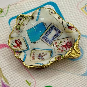 May include: A small, decorative dish made from a natural shell, featuring a gold-painted rim. The interior is decorated with playing card imagery in shades of blue, red, and green. The dish is designed to hold small items.