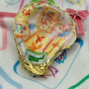 K&ouml;nnte beinhalten: Eine Austernschale mit goldfarbenem Rand, gef&uuml;llt mit einem farbenfrohen Stoffdesign mit floralen und asiatisch inspirierten Motiven. Die Schale liegt auf einem wei&szlig;en Tuch mit einer rosa Schleife.