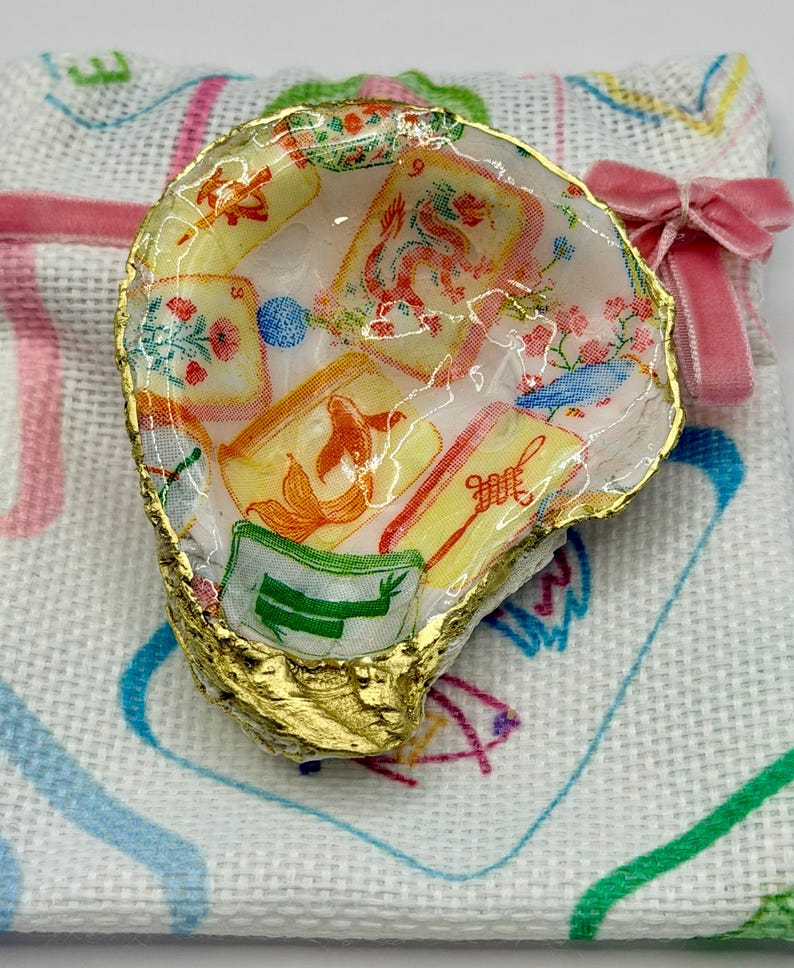 K&ouml;nnte beinhalten: Eine Austernschale mit goldfarbenem Rand, gef&uuml;llt mit einem farbenfrohen Stoffdesign mit floralen und asiatisch inspirierten Motiven. Die Schale liegt auf einem wei&szlig;en Tuch mit einer rosa Schleife.
