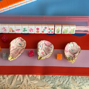 Puede incluir: Tres conchas de ostra decorativas con bordes pintados en dorado, cada una con un patr&oacute;n interior de decoupage de fichas de mahjong. Las conchas est&aacute;n dispuestas junto a varios dados peque&ntilde;os transl&uacute;cidos de color naranja y rosa sobre un tapete de juego colorido.