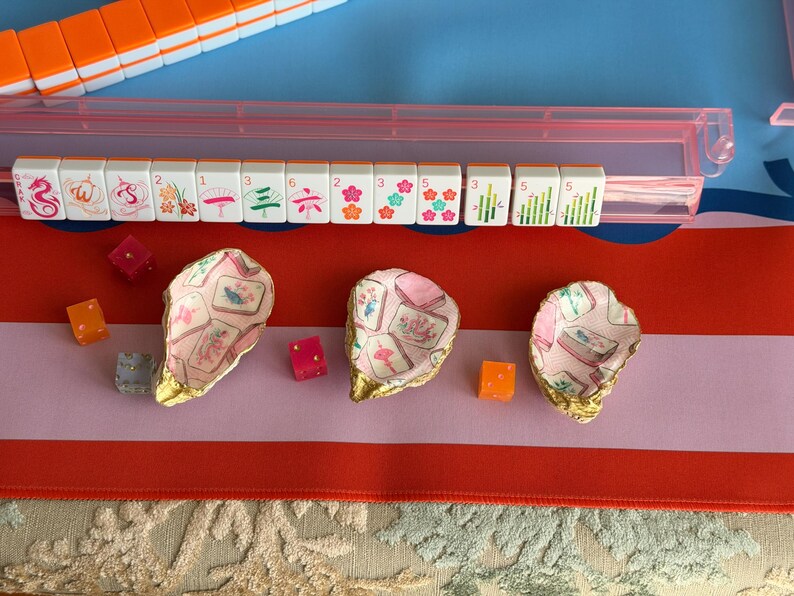 Puede incluir: Tres conchas de ostra decorativas con bordes pintados en dorado, cada una con un patr&oacute;n interior de decoupage de fichas de mahjong. Las conchas est&aacute;n dispuestas junto a varios dados peque&ntilde;os transl&uacute;cidos de color naranja y rosa sobre un tapete de juego colorido.