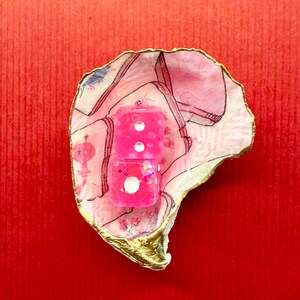 K&ouml;nnte beinhalten: Zwei rosa W&uuml;rfel mit wei&szlig;en Punkten liegen in einer dekorativen, rosa-wei&szlig;-schalenf&ouml;rmigen Schale mit goldenem Rand. Die W&uuml;rfel sind glitzernd und die Schale hat ein Blumenmuster. Der Hintergrund ist einfarbig rot.
