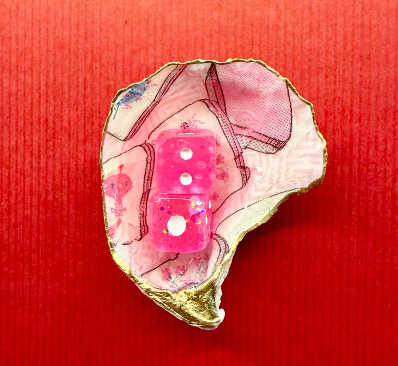 K&ouml;nnte beinhalten: Zwei rosa W&uuml;rfel mit wei&szlig;en Punkten liegen in einer dekorativen, rosa-wei&szlig;-schalenf&ouml;rmigen Schale mit goldenem Rand. Die W&uuml;rfel sind glitzernd und die Schale hat ein Blumenmuster. Der Hintergrund ist einfarbig rot.