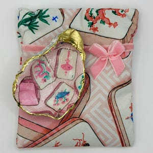 Puede incluir: Una bolsa de tela con estampado de fichas de mahjong en tonos rosa, blanco y verde, acentuada con un lazo de terciopelo rosa. Una concha de ostra decorativa con borde dorado y dise&ntilde;o de ficha de mahjong pintado a mano descansa encima.
