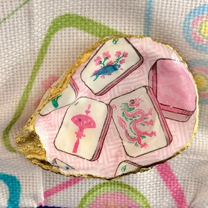 K&ouml;nnte beinhalten: Dekorative Austernschale mit goldfarbenem Rand. Das Innere der Schale ist mit rosa Stoff ausgekleidet, der mit wei&szlig;en, rosa und gr&uuml;nen Mahjong-Kachel-Bildern bedruckt ist. Die Kacheln zeigen V&ouml;gel, Drachen und andere Symbole.
