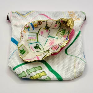 May include: A decorative oyster shell dish with a gold-painted rim, featuring a colorful Mahjong tile design. The dish rests on a small, white fabric pouch with a colorful pattern of pink, green, and blue lines.