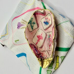 Puede incluir: Una concha de ostra decorativa con borde pintado en oro y un interior de decoupage con un patr&oacute;n de fichas de mahjong en rosa, verde y rojo. La concha descansa sobre una bolsa de tela blanca decorada con dibujos lineales de bamb&uacute;.