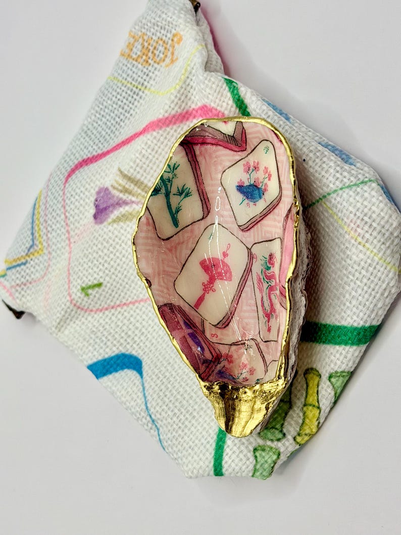 Puede incluir: Una concha de ostra decorativa con borde pintado en oro y un interior de decoupage con un patr&oacute;n de fichas de mahjong en rosa, verde y rojo. La concha descansa sobre una bolsa de tela blanca decorada con dibujos lineales de bamb&uacute;.