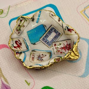 K&ouml;nnte beinhalten: Eine kleine, dekorative Austernschalen-Schale mit goldfarbenem Rand. Das Innere zeigt Spielkarten-Designs in Rot, Blau und Wei&szlig;. Die Schale steht auf einer gemusterten Oberfl&auml;che.