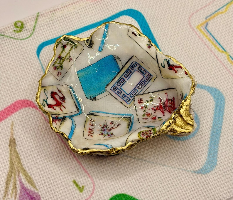 K&ouml;nnte beinhalten: Eine kleine, dekorative Austernschalen-Schale mit goldfarbenem Rand. Das Innere zeigt Spielkarten-Designs in Rot, Blau und Wei&szlig;. Die Schale steht auf einer gemusterten Oberfl&auml;che.