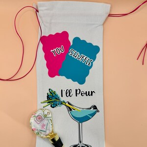 Puede incluir: Una bolsa de vino blanca con el texto "You Shuffle, I'll Pour" y una ilustraci&oacute;n de c&oacute;ctel. Un tap&oacute;n de botella decorativo con un dise&ntilde;o floral y detalles dorados est&aacute; al lado de la bolsa. La bolsa tiene cordones rojos.