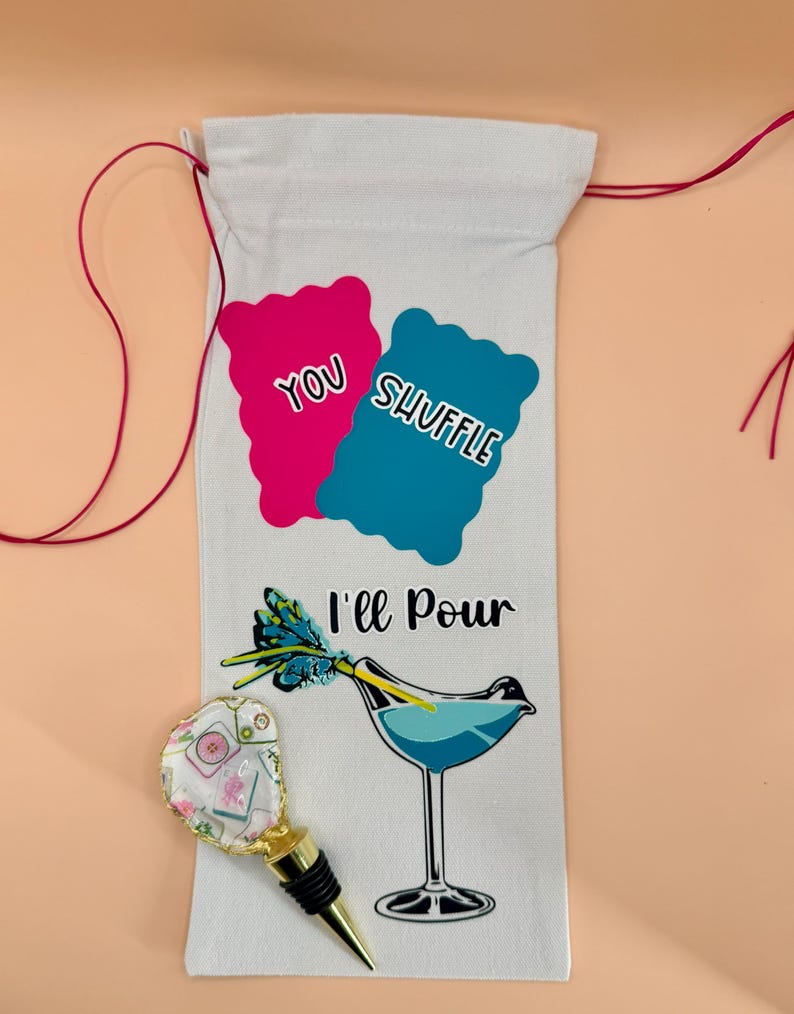 Puede incluir: Una bolsa de vino blanca con el texto "You Shuffle, I'll Pour" y una ilustraci&oacute;n de c&oacute;ctel. Un tap&oacute;n de botella decorativo con un dise&ntilde;o floral y detalles dorados est&aacute; al lado de la bolsa. La bolsa tiene cordones rojos.