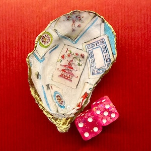 K&ouml;nnte beinhalten: Dekorative Austernschale mit goldenem Rand, bedruckt mit Spielkarten und Pagodenmotiven. Zwei rosa W&uuml;rfel mit wei&szlig;en Punkten liegen neben der Schale, vor rotem Hintergrund.
