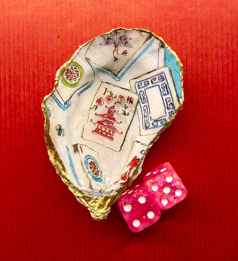 K&ouml;nnte beinhalten: Dekorative Austernschale mit goldenem Rand, bedruckt mit Spielkarten und Pagodenmotiven. Zwei rosa W&uuml;rfel mit wei&szlig;en Punkten liegen neben der Schale, vor rotem Hintergrund.