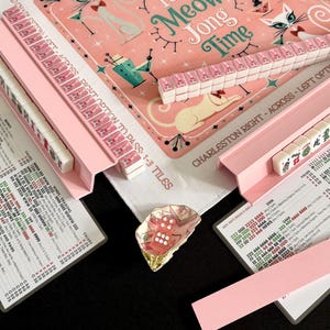 Puede incluir: Un juego de Mahjong con un tapete rosa con ilustraciones de gatos y el texto It's Meow Jong Time. El set incluye soportes rosas, fichas blancas con dise&ntilde;os de gatos rosas, una tarjeta de puntuaci&oacute;n y un dado rosa en un plato de concha.