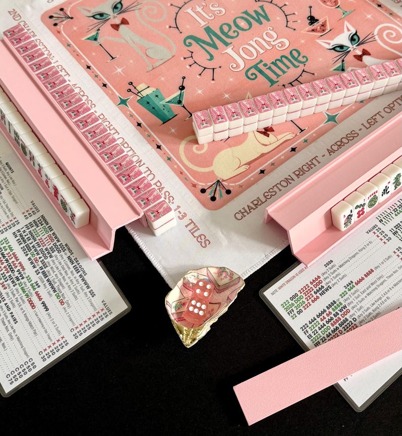 Puede incluir: Un juego de Mahjong con un tapete rosa con ilustraciones de gatos y el texto It's Meow Jong Time. El set incluye soportes rosas, fichas blancas con dise&ntilde;os de gatos rosas, una tarjeta de puntuaci&oacute;n y un dado rosa en un plato de concha.