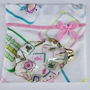 May include: A white fabric pouch with a pink velvet ribbon bow and a colorful mahjong tile print, featuring a white oyster shell trinket dish with a gold-painted rim and a matching mahjong tile pattern resting on top of the pouch.