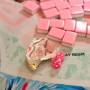 Puede incluir: Una concha de ostra decorativa con borde pintado en oro, con un dise&ntilde;o de fichas de mahjong y un motivo de drag&oacute;n rojo. Junto a la concha hay dos dados peque&ntilde;os de color rosa transl&uacute;cido con puntos blancos, cerca de varias fichas de mahjong rosas.