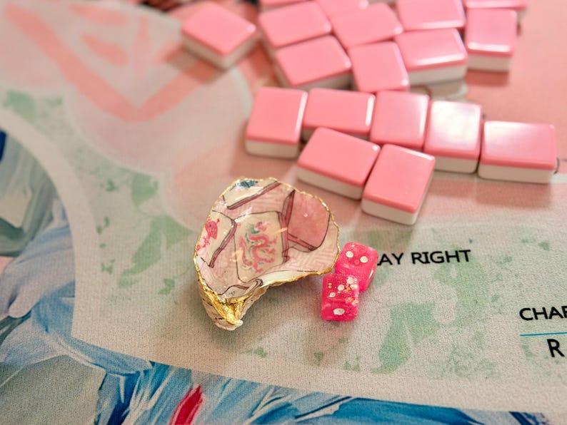 Puede incluir: Una concha de ostra decorativa con borde pintado en oro, con un dise&ntilde;o de fichas de mahjong y un motivo de drag&oacute;n rojo. Junto a la concha hay dos dados peque&ntilde;os de color rosa transl&uacute;cido con puntos blancos, cerca de varias fichas de mahjong rosas.