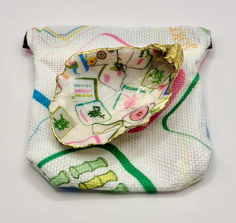 K&ouml;nnte beinhalten: Dekorative Austernschalen-Schale mit goldfarbenem Rand, verziert mit einem farbenfrohen Mahjong-Fliesen-Design. Die Schale liegt auf einem kleinen, wei&szlig;en Stoffbeutel mit einem bunten Muster aus Linien und Bambus.