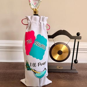 Puede incluir: Bolsa para botella de vino blanco con un dise&ntilde;o rosa y turquesa que dice "You Shuffle, I'll Pour". La bolsa tiene un tap&oacute;n decorativo con un dise&ntilde;o floral. Un peque&ntilde;o gong est&aacute; en el fondo.
