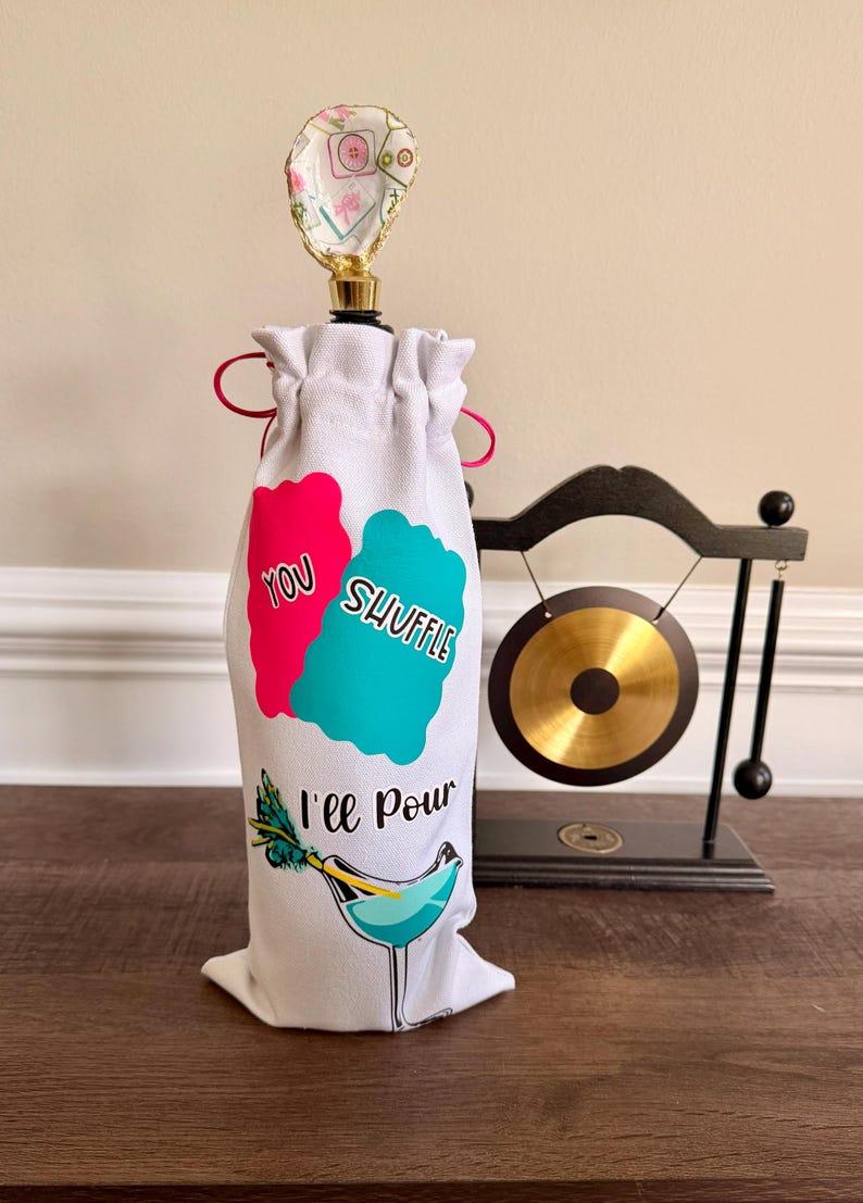 Puede incluir: Bolsa para botella de vino blanco con un dise&ntilde;o rosa y turquesa que dice "You Shuffle, I'll Pour". La bolsa tiene un tap&oacute;n decorativo con un dise&ntilde;o floral. Un peque&ntilde;o gong est&aacute; en el fondo.