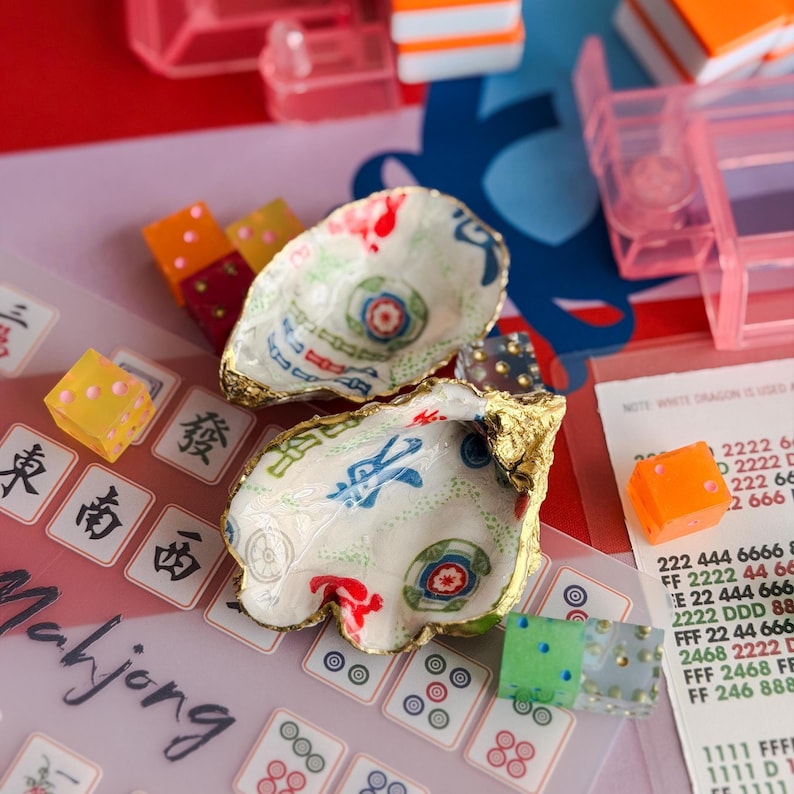 K&ouml;nnte beinhalten: Zwei dekorative Austernschalen mit goldenen R&auml;ndern, die farbenfrohe Muster aufweisen. W&uuml;rfel und Mahjong-Steine sind um die Schalen herum verstreut. Die Schalen haben eine wei&szlig;e Basis mit roten, blauen und gr&uuml;nen Designs. Das Wort "Mahjong" ist sichtbar.